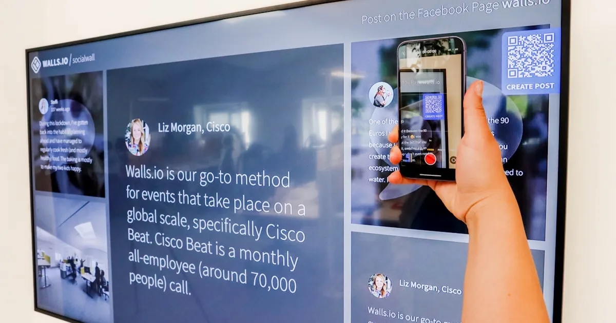 A hand holds a smartphone to scan a QR code displayed on a large digital screen running Walls.io social wall software. The screen displays employee testimonials and event content, showcasing an interactive digital signage solution for corporate engagement.